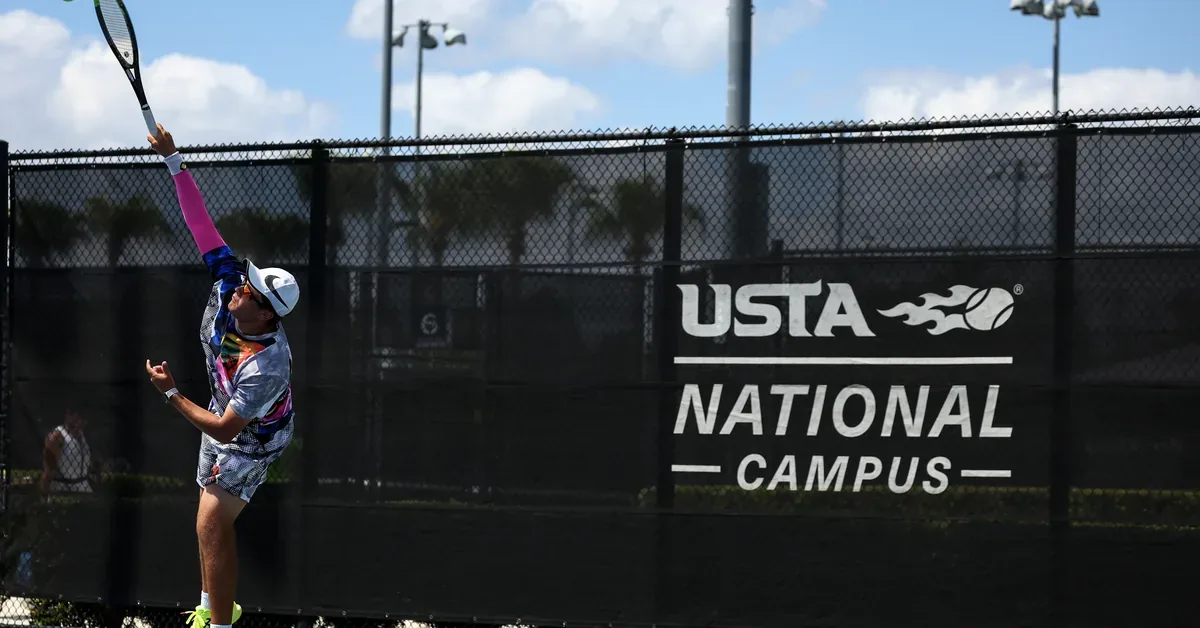 A tennis player executes a powerful serve at the USTA National Campus outdoor courts, surrounded by palm trees under a bright Florida sky.
