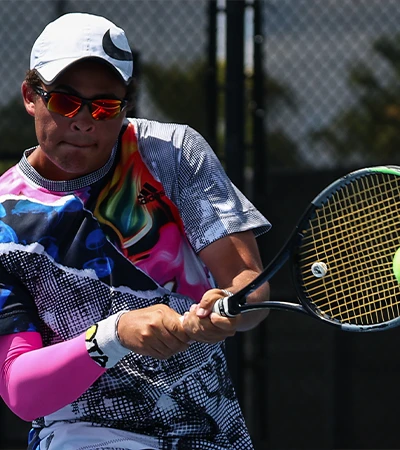 Tennis player in a colorful patterned shirt, white cap, and mirrored sunglasses hitting a two-handed backhand, tennis ball on the racquet strings, chain-link fence in the background.