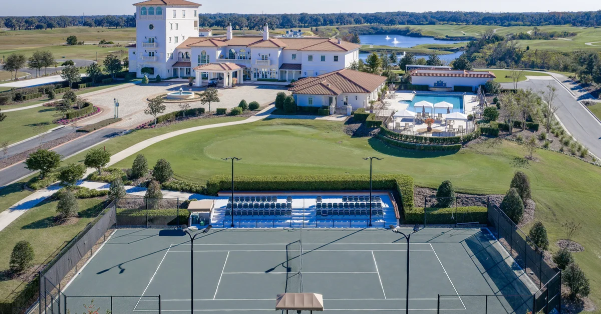 Aerial view of a large Mediterranean-style clubhouse next to a swimming pool, putting green, and tennis court with stadium seating, bordered by a lake with fountains and a golf course.