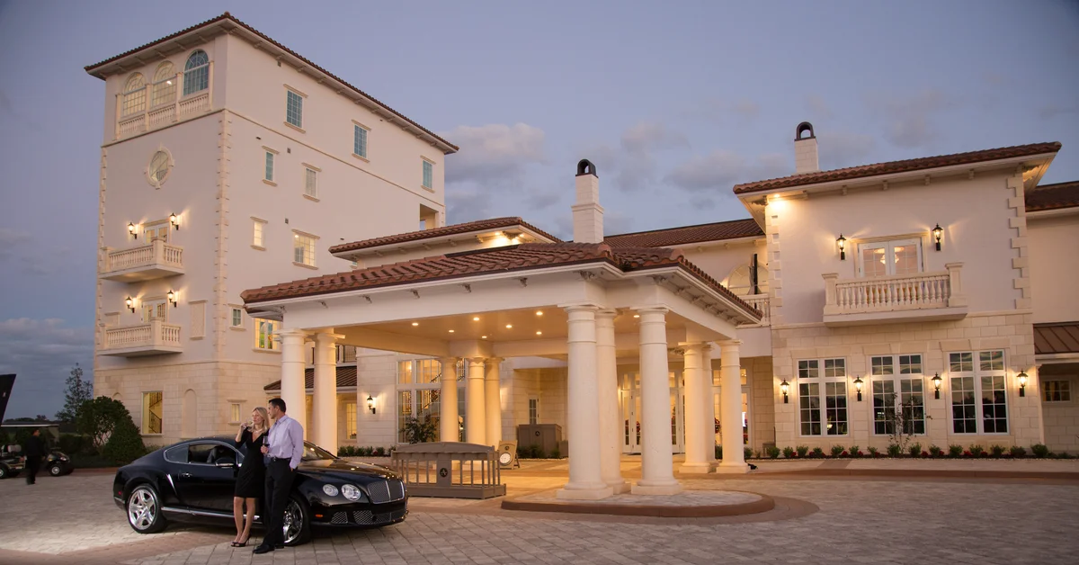 Man and woman in evening attire standing beside a black luxury car under a lit column entrance of a cream-colored Mediterranean clubhouse at dusk.