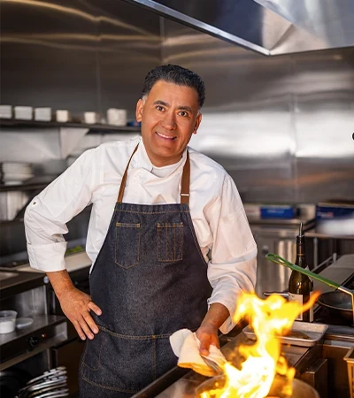 Executive Chef Hugo Marroguin in a white chef coat and denim apron, smiling while flambeing a pan with visible flames in a stainless steel restaurant kitchen.